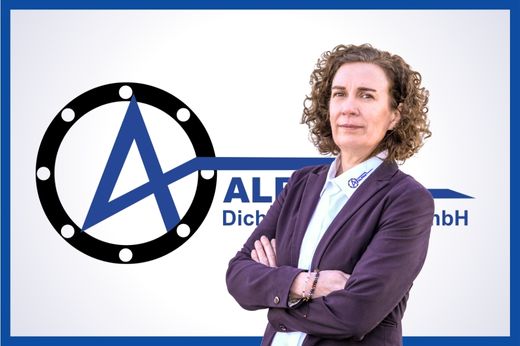 Produktionsleitung Ina Löschmann mit weißem Hemd und dunkelblauem Blazer mit verschränkten Armen vor dem blauen Alpha-Logo auf weißem Hintergrund.
