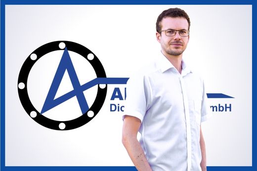 Einkäufer Toni Kaminsky mit weißem Hemd in legerer Haltung vor dem blauen Alpha-Logo auf weißem Hintergrund.